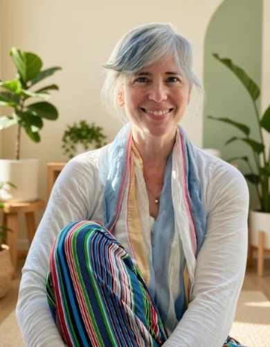 Stacy Joslin, smiling, seated cross-legged in a bright room with plants